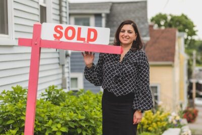 A smiling real estate agent stands next to a "SOLD" sign in front of a house